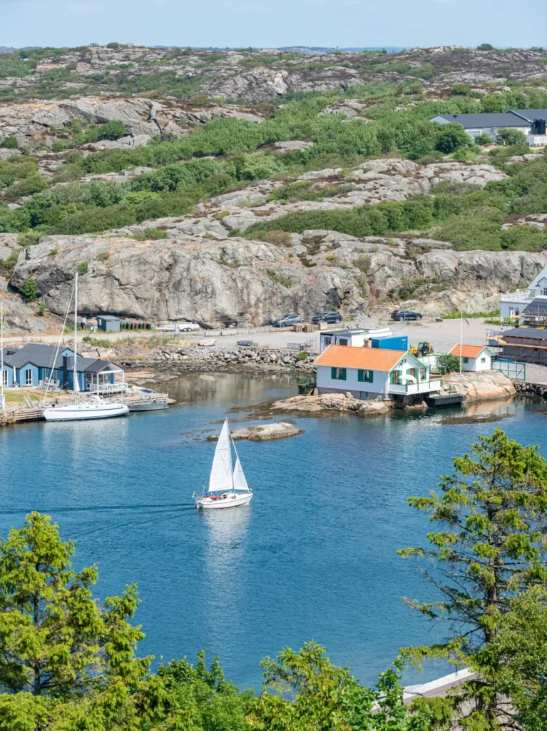 En segelbåt glider genom en vacker skärgårdsmiljö med klippor, grönska och små hus vid vattnet. Perfekt exempel på en fastighet i behov av tillsyn.