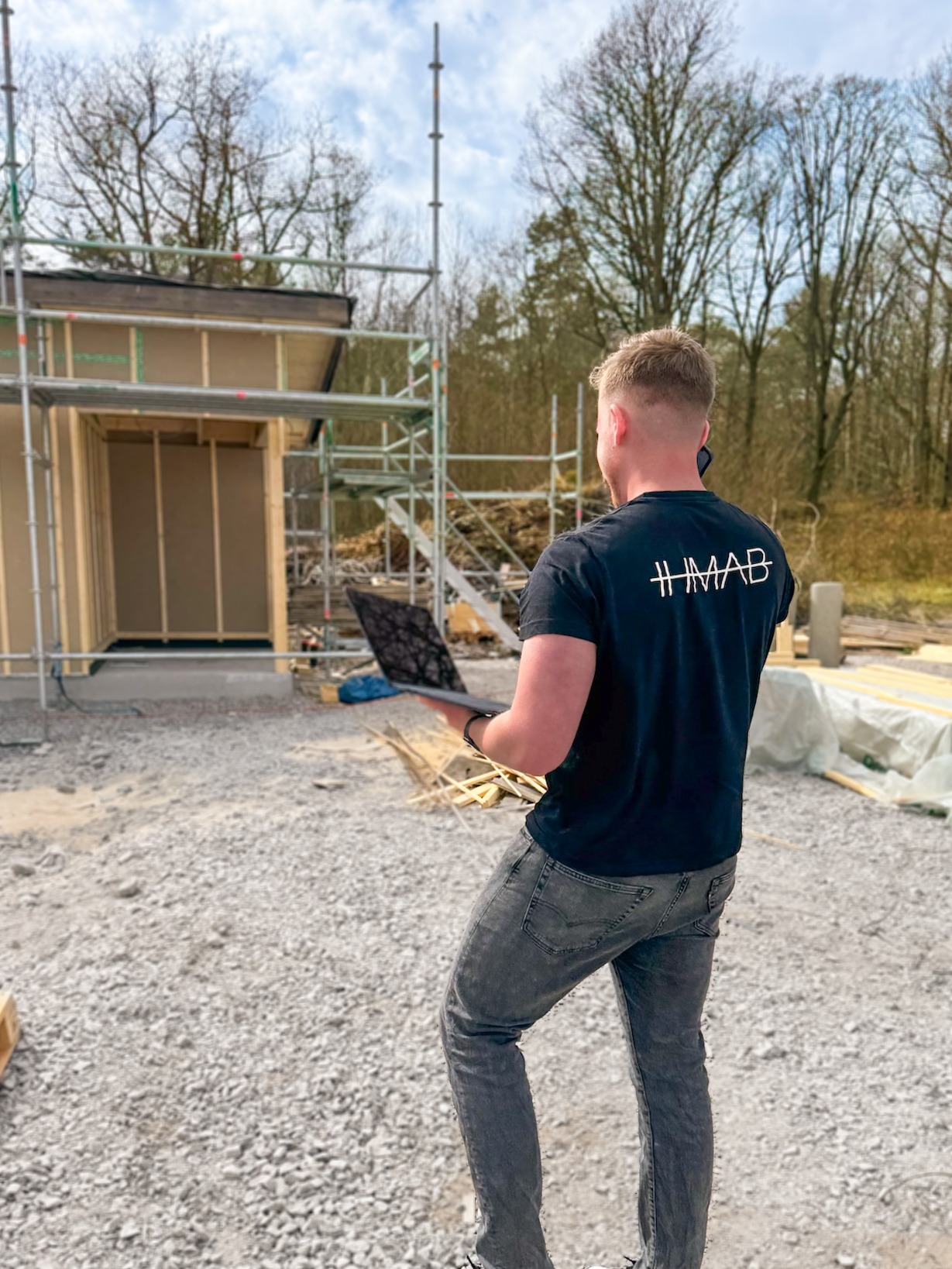 Man med HMAB-tröja inspekterar byggarbetsplats med bärbar dator i handen, framför ett hus under uppbyggnad med byggställningar – omgiven av träd och byggmaterial.